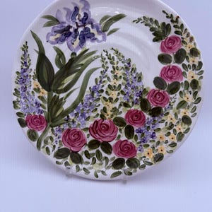 May include: A round white ceramic plate featuring a hand-painted floral design. The pattern includes large pink roses, clusters of small purple and yellow wildflowers, and green leaves arranged in a curved, asymmetrical composition across the surface.