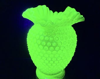 May include: A bright neon green glowing vase featuring a textured hobnail pattern across its rounded body and a wide, ruffled, scalloped rim. The glass object emits a vivid fluorescent light against a dark background, highlighting its unique surface.