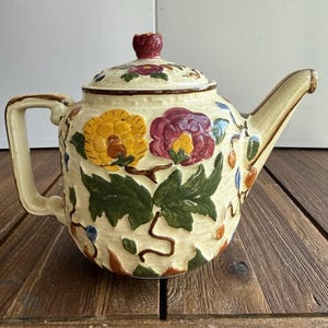 May include: A cream-coloured ceramic teapot featuring a raised, embossed floral design with large yellow and purple flowers, green leaves, and brown stems. The teapot has a matching lid with a small knob, a curved handle, and a tapered spout.
