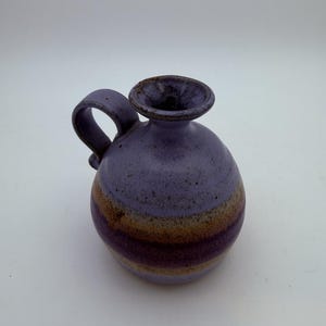 May include: A small, round ceramic jug with a single handle and a flared rim. The body features a speckled lavender glaze on the top and bottom, with a wide, horizontal band of earthy brown and tan tones encircling the center of the vessel.