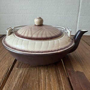 May include: A ceramic teapot with a dark brown base and a cream-coloured lid featuring a radial leaf-like pattern and dark brown edges. The teapot includes a metal swing handle attached to two side loops and a dark brown spout on the right side.