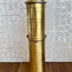 May include: A tall, cylindrical brass pepper mill featuring horizontal engraved lines along its body, a metallic top cap, and a dark, weathered iron hand crank mechanism attached to the top for manual grinding of spices or peppercorns.
