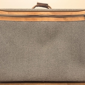 May include: A rectangular suitcase featuring a textured grey and brown tweed fabric body with tan leather trim along the edges and a horizontal zipped pocket across the top. The luggage includes a sturdy leather handle attached to the top centre.