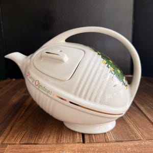 May include: A white ceramic teapot with a curved handle and a spout. The body features vertical ribbed detailing, a gold trim line, and a colourful Christmas tree illustration. The text Merry Christmas is printed in red script on the side.
