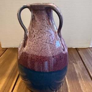 May include: A ceramic vase featuring two curved handles and a multi-toned glaze. The top section is a speckled light mauve, transitioning into a solid deep burgundy band in the middle, and finishing with a dark navy blue base at the bottom.
