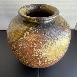 May include: A round ceramic vase with a wide opening and a thick, rolled rim. The exterior features a textured, mottled glaze in shades of earthy brown, rust orange, and olive green, with a glossy, dripped finish near the top rim of the vessel.