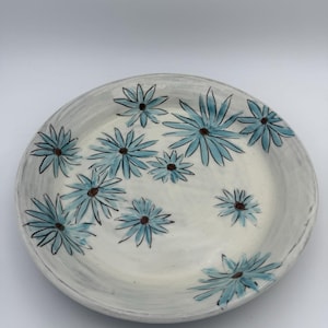 May include: A round ceramic plate with a white base and a hand-painted pattern of light blue daisy-like flowers. Each flower features thin, elongated petals radiating from a small, dark brown center. The surface has visible brushstroke textures.