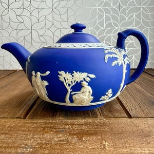 May include: A cobalt blue teapot with a matte finish, featuring white relief ornamentation of classical figures and trees. The teapot includes a matching lid with a small knob, a curved spout, and a sturdy handle, showcasing intricate detailing.