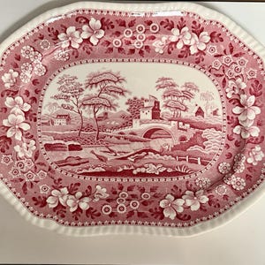 May include: An oval serving platter with a scalloped edge, featuring a red and white transferware pattern. The center depicts a landscape with a stone bridge, buildings, and trees, while the border displays a floral motif of blossoms and leaves.