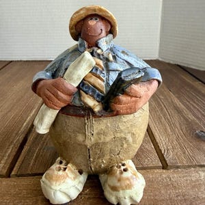 May include: A ceramic figurine of a round-bodied character wearing a tan hat, a blue collared shirt, and a striped tie. The figure holds a rolled white paper in one hand and a black rectangular object in the other, wearing tan pants and white shoes.