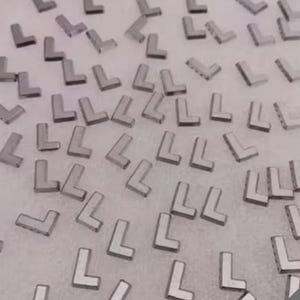 Pode incluir: Uma coleção de pequenas peças metálicas prateadas em forma de L espalhadas sobre uma superfície clara. As peças parecem uniformes em tamanho e forma, com uma qualidade reflexiva.