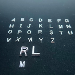 Peut inclure: Une collection de lettres individuelles en métal de couleur argentée disposées en rangées, présentant l'alphabet complet de A à Z en majuscules, avec des lettres R, L et M supplémentaires positionnées sous la séquence principale.