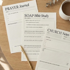 May include: Three white paper journals with black text, including "Prayer Journal", "SOAP Bible Study", and "Church Notes", are arranged on a light wood surface. A gold pen lies across the pages, and a white mug with a dark beverage is in the background.