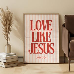 Puede incluir: Un estampado enmarcado con las palabras "LOVE LIKE JESUS" en letras rojas y en negrita. El fondo tiene rayas verticales rosas y blancas. El texto "JOHN 13:34" está en la parte inferior. El estampado está en una habitación con un jarrón de flores secas y un sofá marrón.