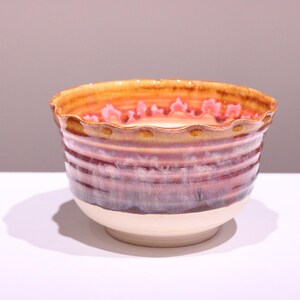 Puede incluir: Un cuenco de cerámica con borde ondulado. El cuenco presenta una base color crema, que pasa a un cuerpo morado y marrón con estrías horizontales. El borde es de color naranja cálido con detalles en rosa. El cuenco es probablemente hecho a mano.