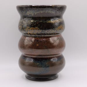 May include: A cylindrical ceramic vase with a stacked, bulbous silhouette. The surface features a glossy, mottled glaze in shades of deep brown, black, and hints of metallic bronze and blue, creating a textured, multi-layered appearance.