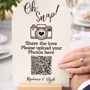 May include: A cream-colored wedding sign with the words "Oh Snap!" and a camera graphic with a heart in the lens. The sign includes a QR code and the text "Share the love Please upload your Photos here".