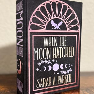 May include: A hardcover book titled "When The Moon Hatched" by Sarah A. Parker. The book has a black cover with pink and white lettering and design elements, including a bat silhouette and moon phases.