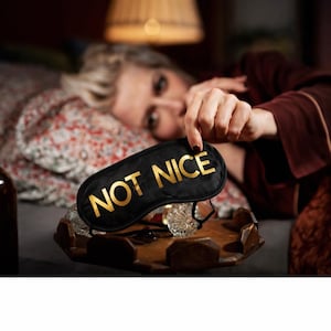 Op de afbeelding: Zwart slaapmasker met gouden tekst "NOT NICE". Het masker wordt omhoog gehouden voor een persoon die in bed ligt. Het bed heeft een bloemenpatroon dekbed. Een houten dienblad met glazen voorwerpen staat op de voorgrond.