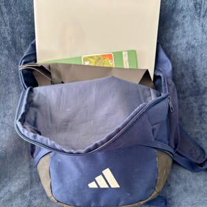 May include: A navy blue backpack with a white Adidas logo, holding a laptop and a green and orange book. The backpack has a black bottom and a mesh side pocket. The bag is open, revealing its contents.