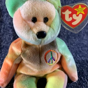 May include: A colorful plush bear toy with a peace sign on its chest. The bear has a rainbow tie-dye pattern with green, orange, and yellow hues. It has black eyes and a black nose. A red heart-shaped tag is attached.