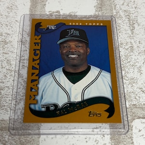 May include: A 2002 Topps baseball card featuring a portrait of manager Hal McRae wearing a white Tampa Bay Rays jersey and a black cap. The card has a gold border with the text MANAGER printed vertically on the left and the name Hal McRae below.