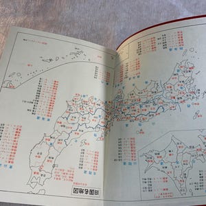May include: Open book with a detailed map of Japan, featuring red and blue text labels and outlines. The map is divided into regions and prefectures, with a smaller inset map. The book has a red cover.