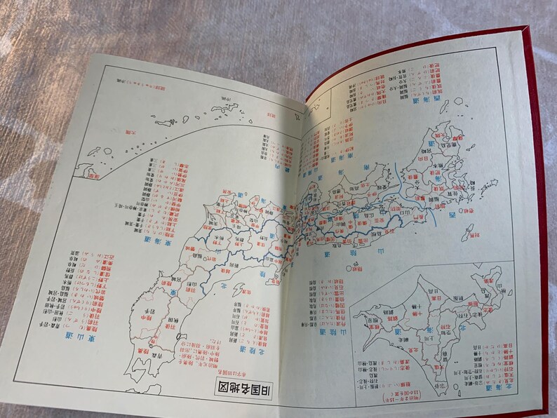 May include: Open book with a detailed map of Japan, featuring red and blue text labels and outlines. The map is divided into regions and prefectures, with a smaller inset map. The book has a red cover.