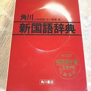 May include: A red book with Japanese text. The title "新国語辞典" is prominently displayed in black. Additional text includes the publisher's name and a gold seal with more Japanese characters. The book has a white and red design.