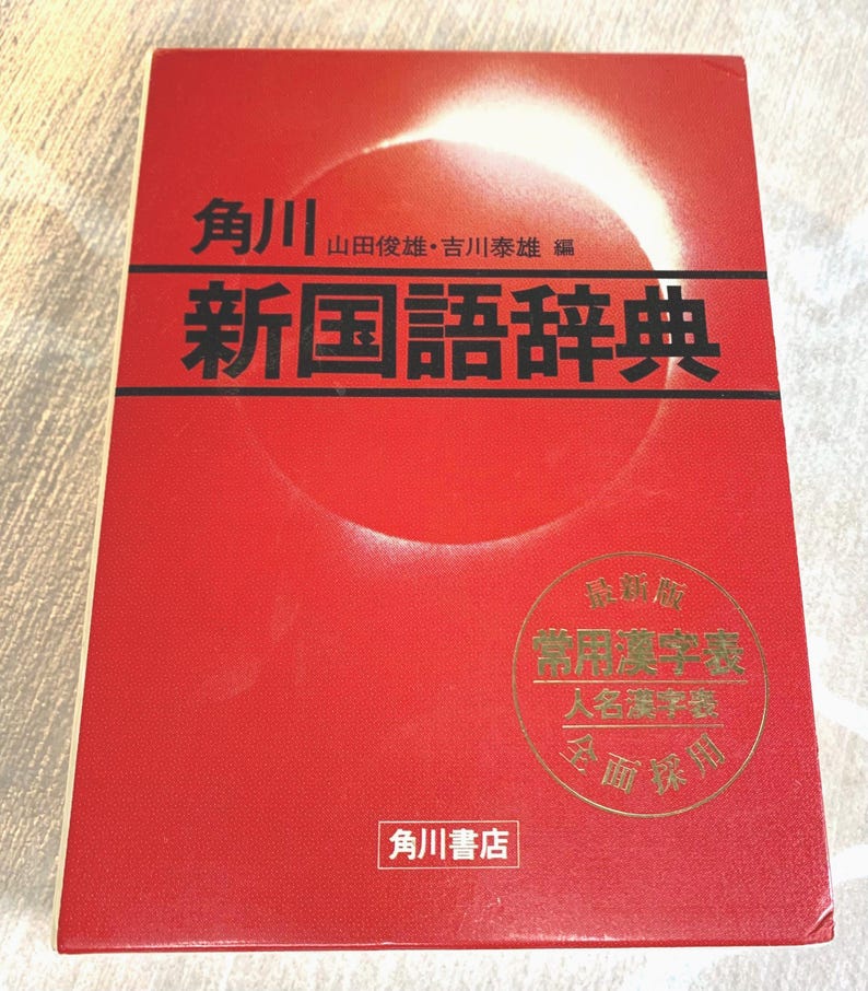May include: A red book with Japanese text. The title "新国語辞典" is prominently displayed in black. Additional text includes the publisher's name and a gold seal with more Japanese characters. The book has a white and red design.