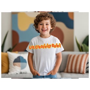 May include: A white t-shirt with the text "Let Creativity Rule!" in orange and yellow. The shirt is worn by a smiling child. The child is standing in front of a couch with decorative pillows.