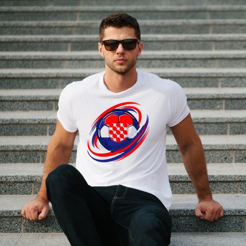May include: White t-shirt featuring a soccer ball design with the Croatian flag colors: red, white, and blue. The soccer ball is surrounded by red and blue curved lines. The shirt is a crew neck style.