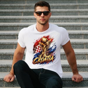May include: White t-shirt featuring a golden trophy with a soccer ball, the Croatian flag, and the word "CROATIA" in gold. The design is set against a backdrop of red, white, and blue brushstrokes.
