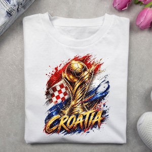 May include: White t-shirt featuring a golden trophy with a soccer ball on top, set against a backdrop of red, white, and blue paint strokes and the word "CROATIA" in gold. The design incorporates the Croatian flag.
