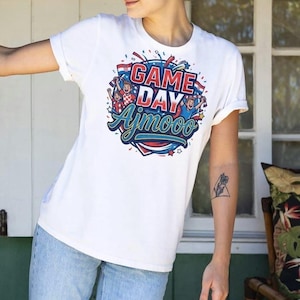 May include: White t-shirt with a colorful graphic that says "GAME DAY Ajmooo". The design features cheering figures, confetti, and a flag. The shirt has short sleeves and a crew neck.