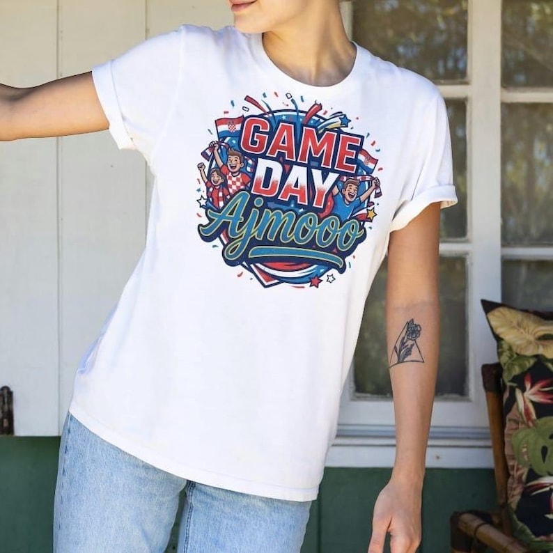 May include: White t-shirt with a colorful graphic that says "GAME DAY Ajmooo". The design features cheering figures, confetti, and a flag. The shirt has short sleeves and a crew neck.