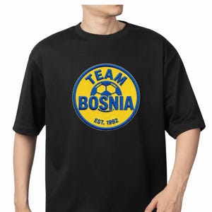 May include: Black t-shirt with a circular graphic featuring the words "TEAM BOSNIA" in blue and a soccer ball design on a yellow background. The text "EST. 1992" is below the main text. The shirt is short-sleeved.