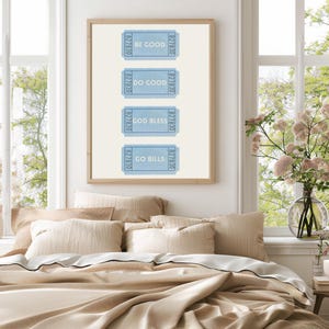 Peut inclure: Une affiche encadrée avec quatre billets bleus portant les inscriptions « BE GOOD », « DO GOOD », « GOD BLESS » et « GO BILLS ». L'œuvre est au-dessus d'un lit avec une literie beige, sur fond de fenêtres et de lumière naturelle.