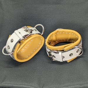 Puede incluir: Dos esposas de cuero para muñeca. Cada una tiene un interior marrón y un exterior blanco con herrajes metálicos plateados. Las esposas se aseguran con hebillas y tienen anillos metálicos.