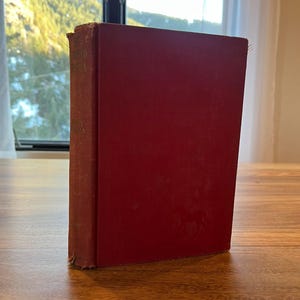 May include: A vintage, hardcover book with a deep red cover. The spine shows signs of wear, and the pages are slightly aged. The book is standing upright on a wooden surface, with a window and mountain view in the background.