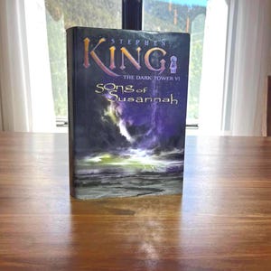 Könnte beinhalten: Ein Hardcover-Buch mit dem Titel "Song of Susannah" von Stephen King, aus der Reihe "The Dark Tower VI". Das Cover zeigt eine dunkle, dramatische Szene mit einem stürmischen Himmel und Blitzen über dem Wasser.
