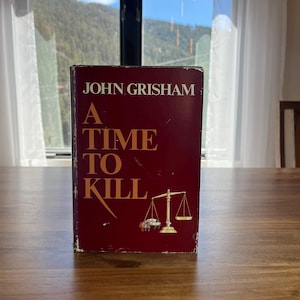 May include: A hardcover book titled "A Time to Kill" by John Grisham. The cover is a deep red with gold lettering and a gold scale illustration. The book is standing on a wooden table in front of a window.