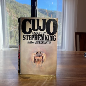 Puede incluir: Un libro de tapa dura titulado "CUJO" de Stephen King. La portada del libro presenta el título en letras negras en negrita, con el nombre del autor debajo. La portada muestra la cara de un perro gruñendo.