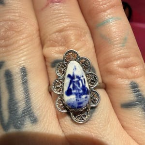 May include: A silver ring with a white and blue porcelain centerpiece. The porcelain has a blue landscape design. The ring has a filigree design around the porcelain. The ring is on a finger with tattoos.