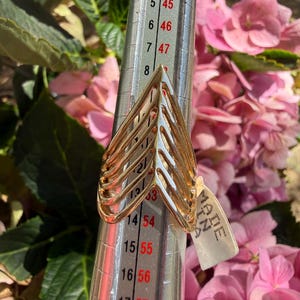 May include: A gold-toned chevron ring with a multi-band design, positioned on a metal ring mandrel. The mandrel displays numerical sizing markings, with the ring resting near the 13 mark. A small white tag labeled MADE IN ITALY is attached.