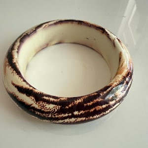 May include: A wide, round bangle bracelet with a cream-colored interior and a dark brown and tan patterned exterior. The bracelet has a glossy finish and a thick, solid construction. The pattern resembles a tiger stripe design.