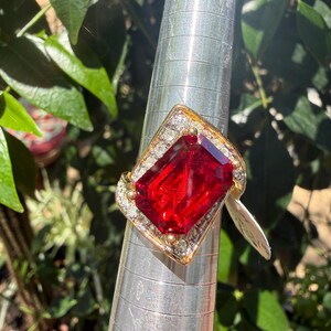 May include: A gold-toned ring featuring a large, rectangular, faceted red stone at the center, surrounded by a border of small, clear, sparkling stones set in a geometric frame, displayed on a silver-colored metal ring mandrel.