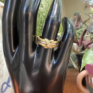 May include: A gold-toned ring with a diamond-like cluster at the center, set on a black display hand. The ring features a layered design with silver-toned bands. The background includes greenery and a decorative mug.