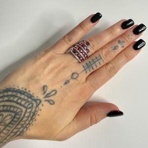 May include: A silver ring with a heart-shaped design. The ring features multiple oval-cut red gemstones and is surrounded by smaller, clear gemstones. The ring is on a hand with black nail polish.