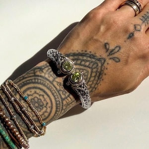 May include: Silver cuff bracelet with an ornate filigree design and two round, light green gemstones. The bracelet is worn on a wrist with multiple other bracelets and tattoos. The gemstones are a bright, spring green colour.